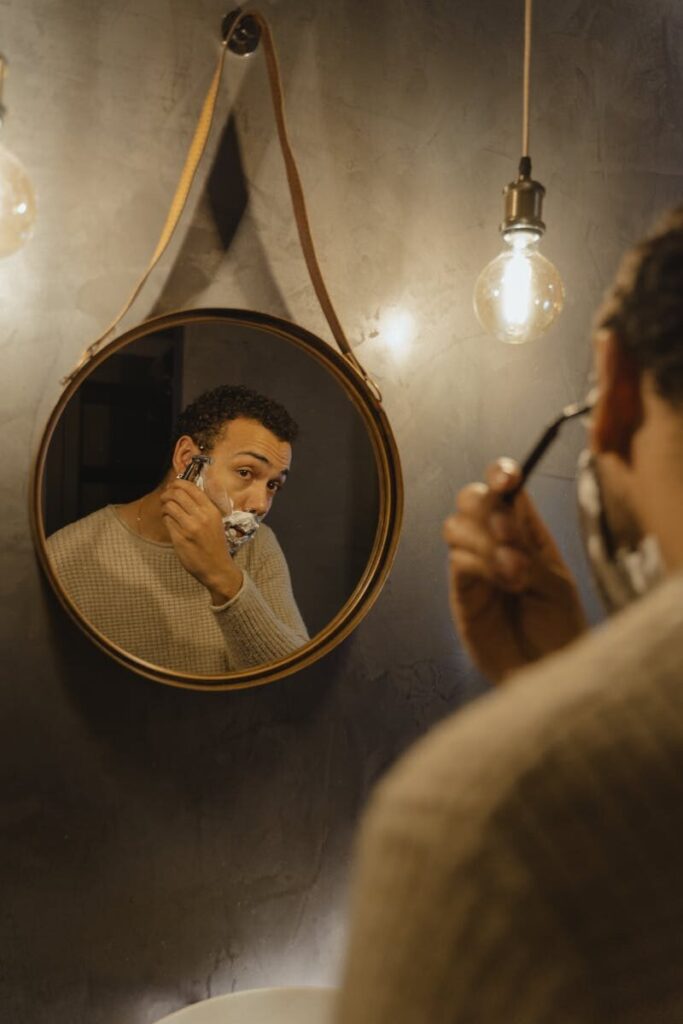 An adult man grooming his beard using a razor in front of a round mirror under warm lighting.