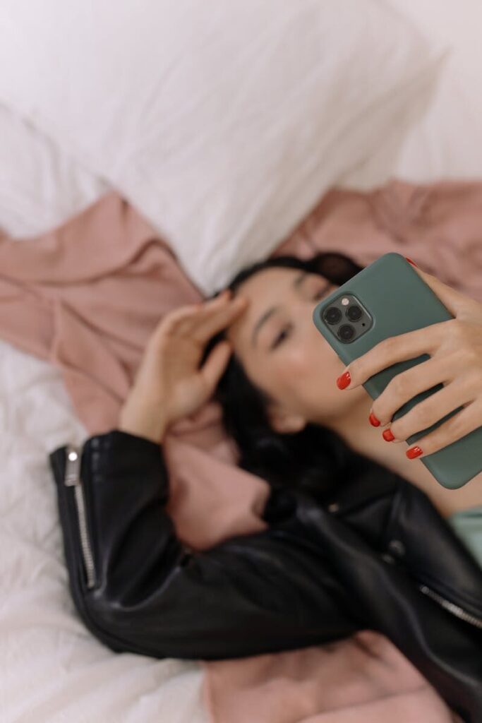 A woman lying on a bed taking a selfie with her smartphone, wearing a leather jacket.