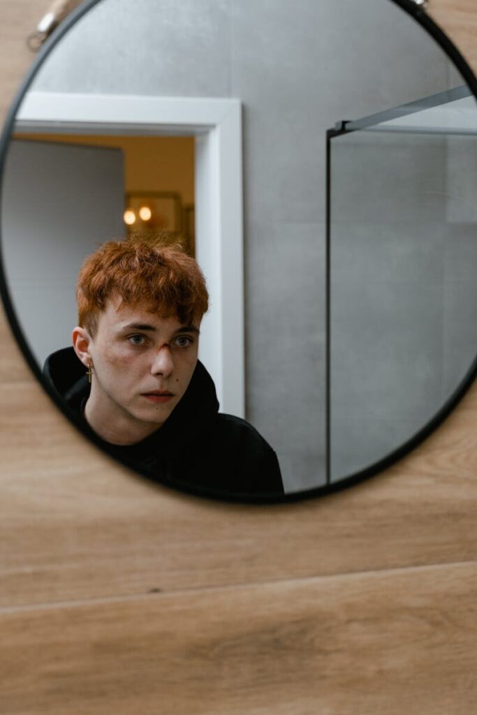 Pensive teenager with red hair looking at his reflection in a bathroom mirror indoors.